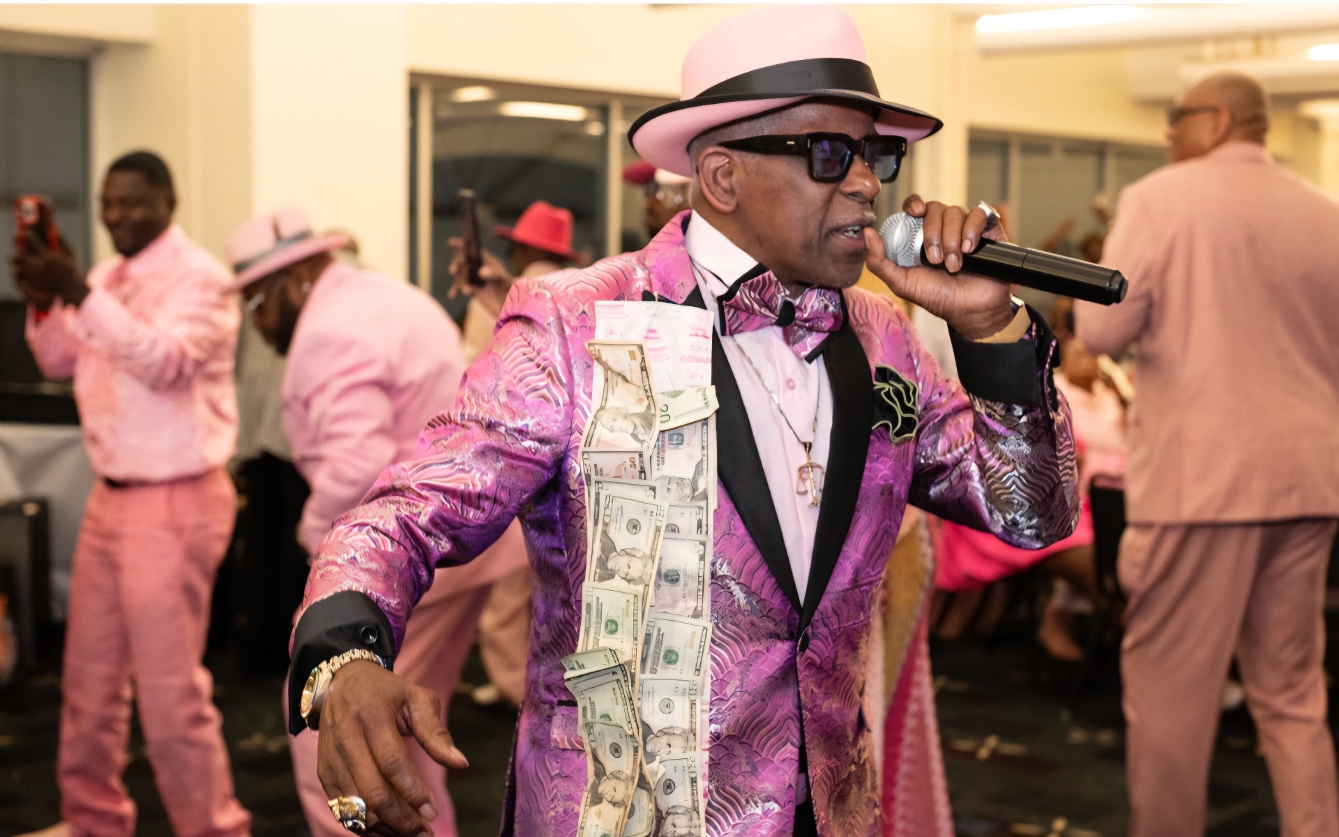 Fast Freddy Anderson on the microphone in a pink brocade jacket and pink fedora at his 80th birthday celebration.
