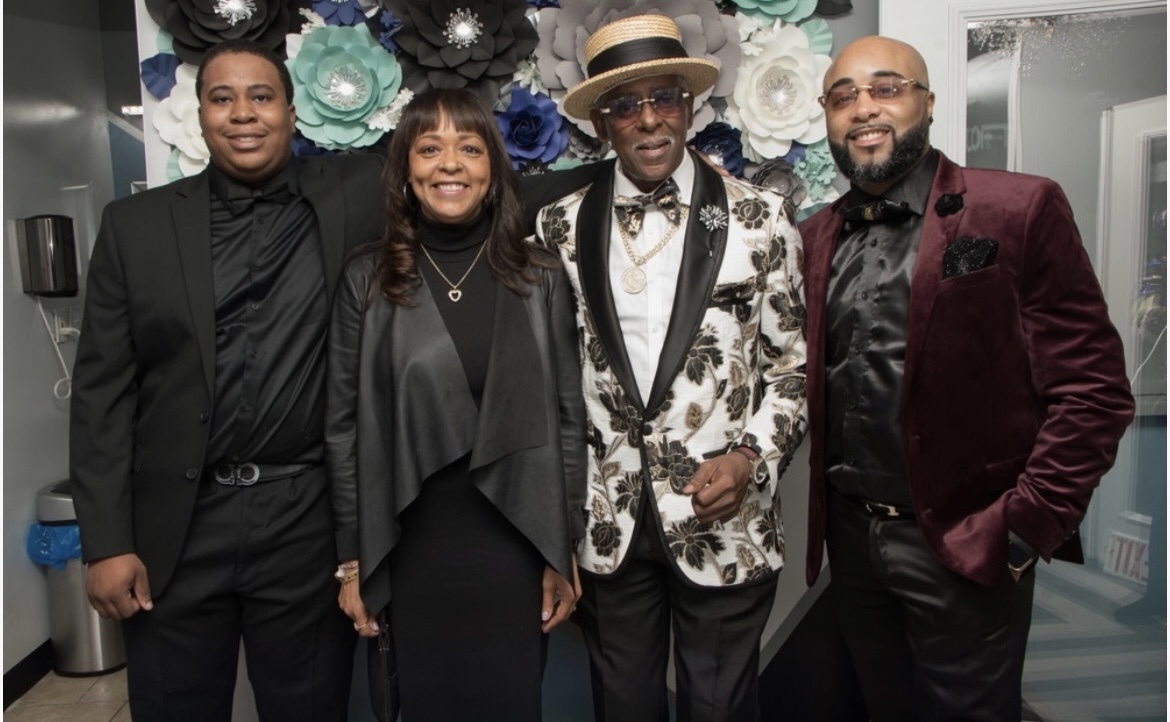 Fast Freddy Anderson in a black and gold floral dinner jacket and straw boater hat with three formally dressed family members at an event.