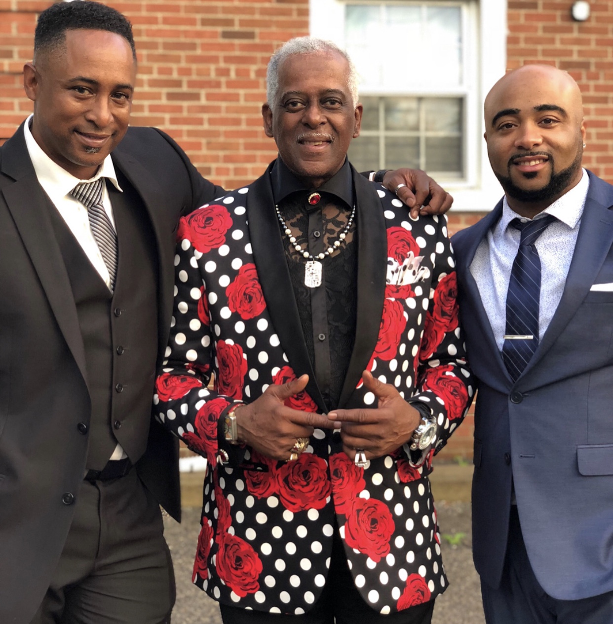 Fast Freddy Anderson in a red rose polka dot jacket standing with two suited family members outside a Detroit home.