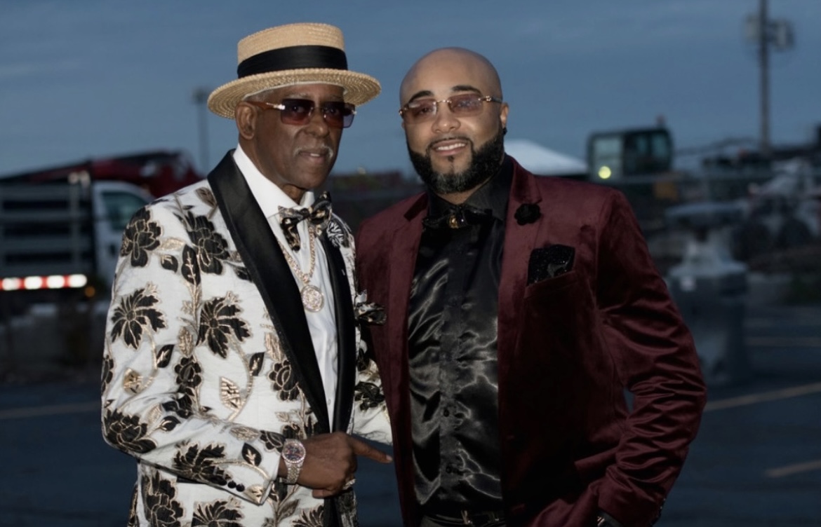 Fast Freddy Anderson in a black and gold floral jacket posing with a friend in a burgundy velvet blazer.