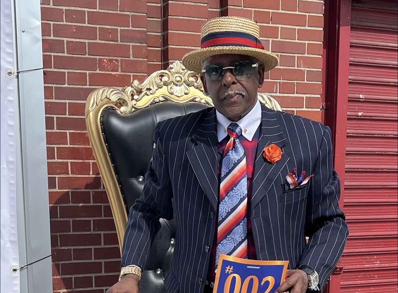 Fast Freddy Anderson seated in a gold throne chair wearing a pinstripe suit and straw boater hat.