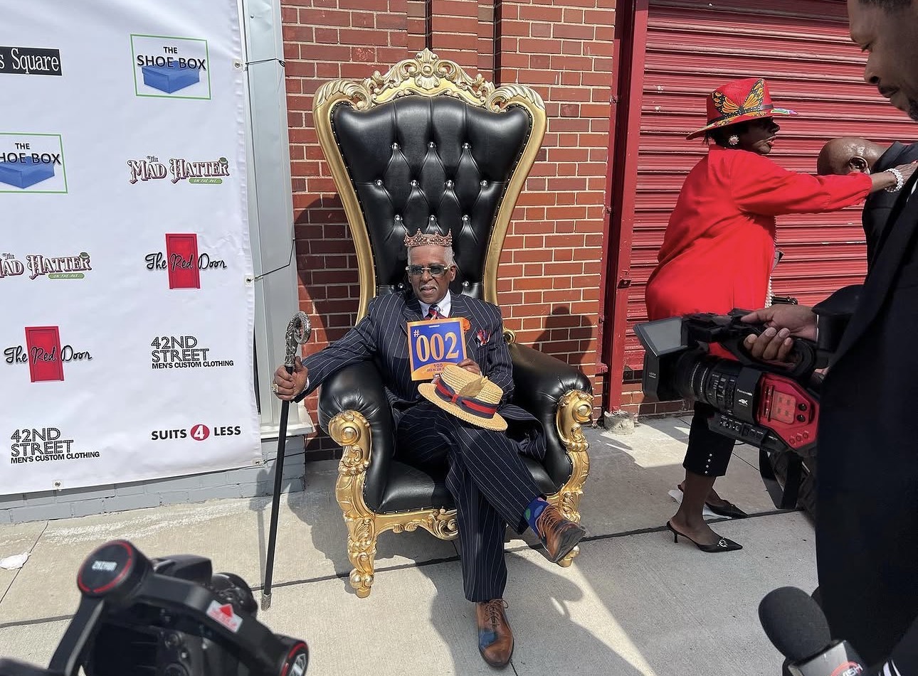 Fast Freddy Anderson crowned and seated on a gold throne on a Detroit sidewalk while news cameras film.